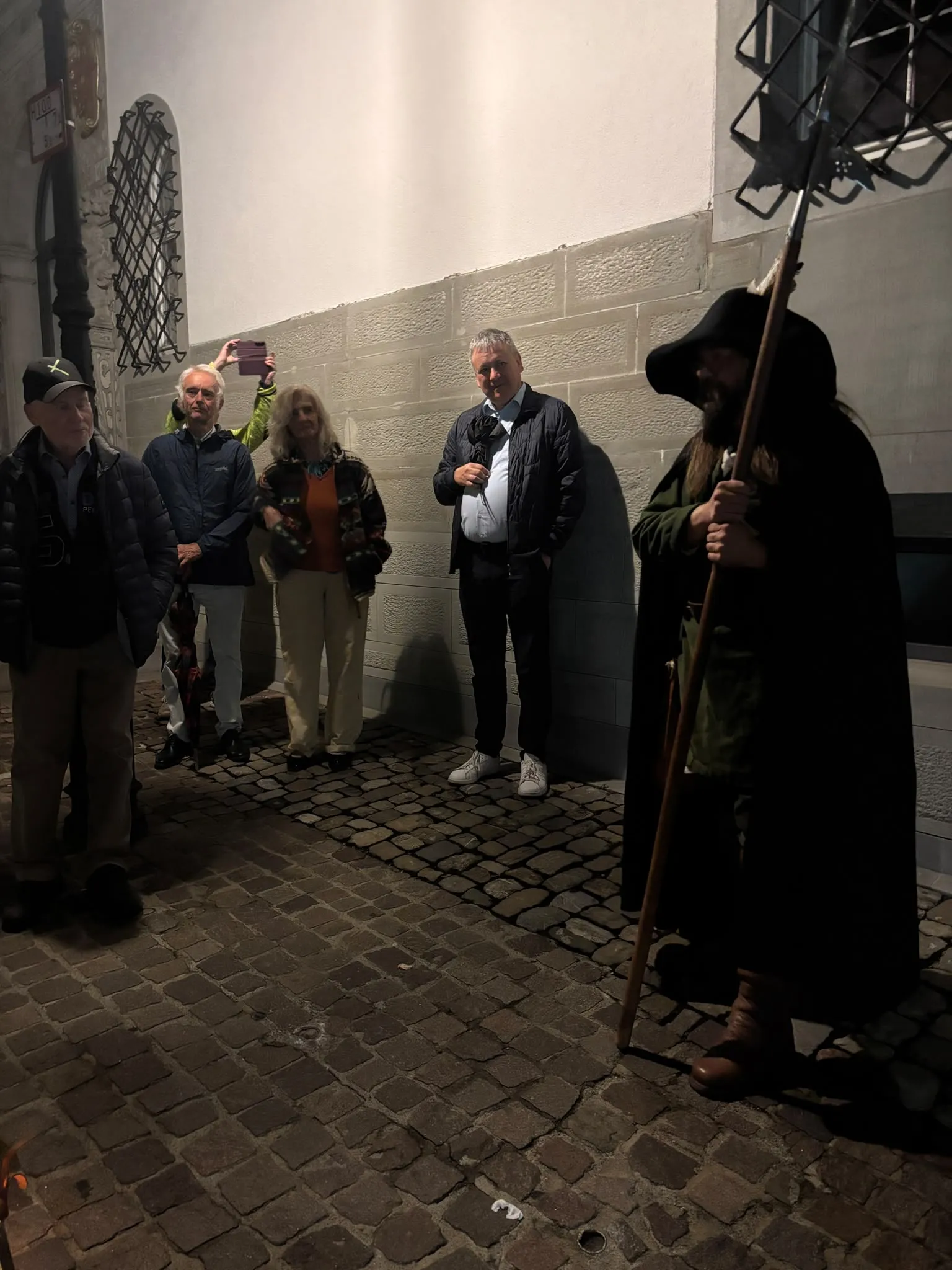 Teilnehmende hören dem Nachtwächter in einer beleuchteten Gasse mit Kopfsteinpflaster zu
